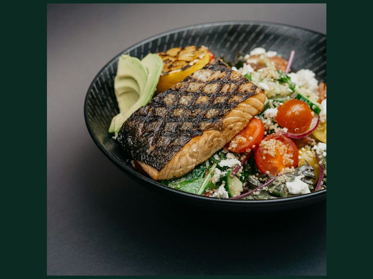 A grilled salmon power bowl — salmon over quinoa, greens, tomato, avocado and feta in a dark bowl — a real meal, protein at the center